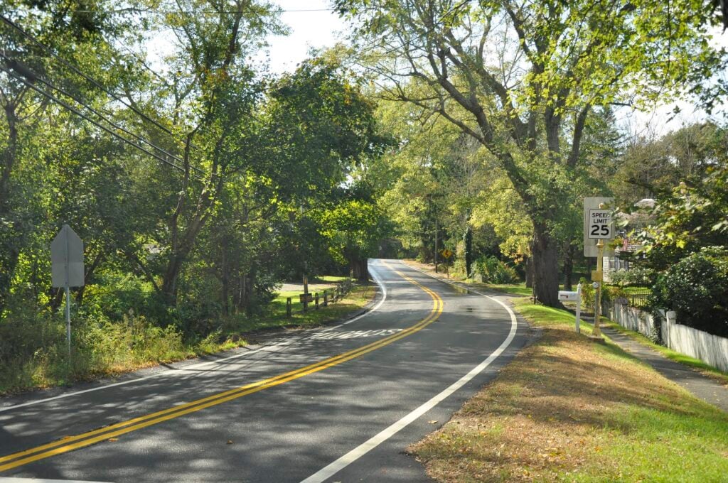 The winding paved road of Route 6A on Cape Cod, better known as Old King's Highway, flanked by trees on either side.