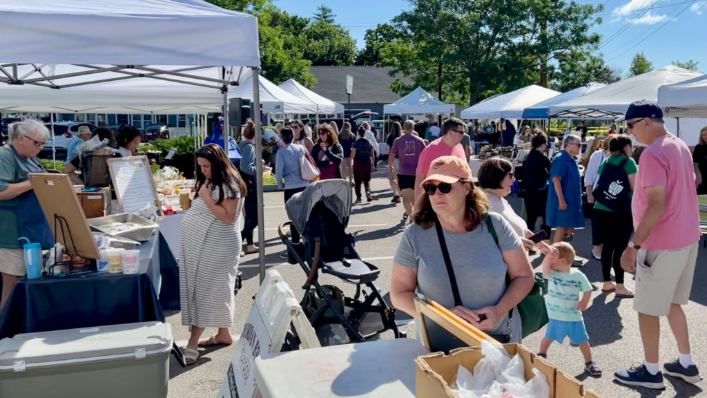 People browse vendor stalls at an outdoor farmers market on a sunny day. Discover Massachusetts Farmers Markets as tents and tables display goods, with families and children mingling in the crowd and a woman in sunglasses walking by.