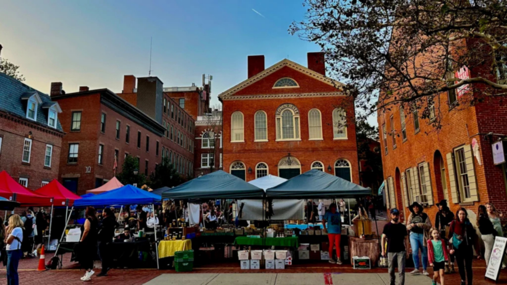 Discover Massachusetts Farmers Markets at this vibrant outdoor market, where colorful tents and vendors line a brick-paved street in front of historic red brick buildings, inviting people to browse under a bright blue sky.