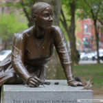 A bronze statue of a woman kneeling and writing on a stone, with a small bird perched on the edge. Lush green trees and blurred buildings appear in the background. A photo credit reads “Photo credit Pat Greenhouse.”.