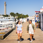 A smiling couple walks along a sunny marina boardwalk, with boats docked on one side and shops on the other. A tall stone tower rises in the background under a clear blue sky.