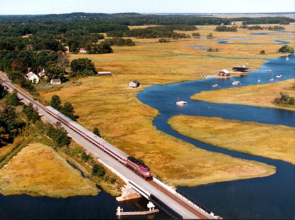 Aerial view of a train crossing a bridge over winding waterways in a marshy Massachusetts landscape, with scattered houses, trees, and grassy fields—perfect for those looking to explore without a car under a clear sky.