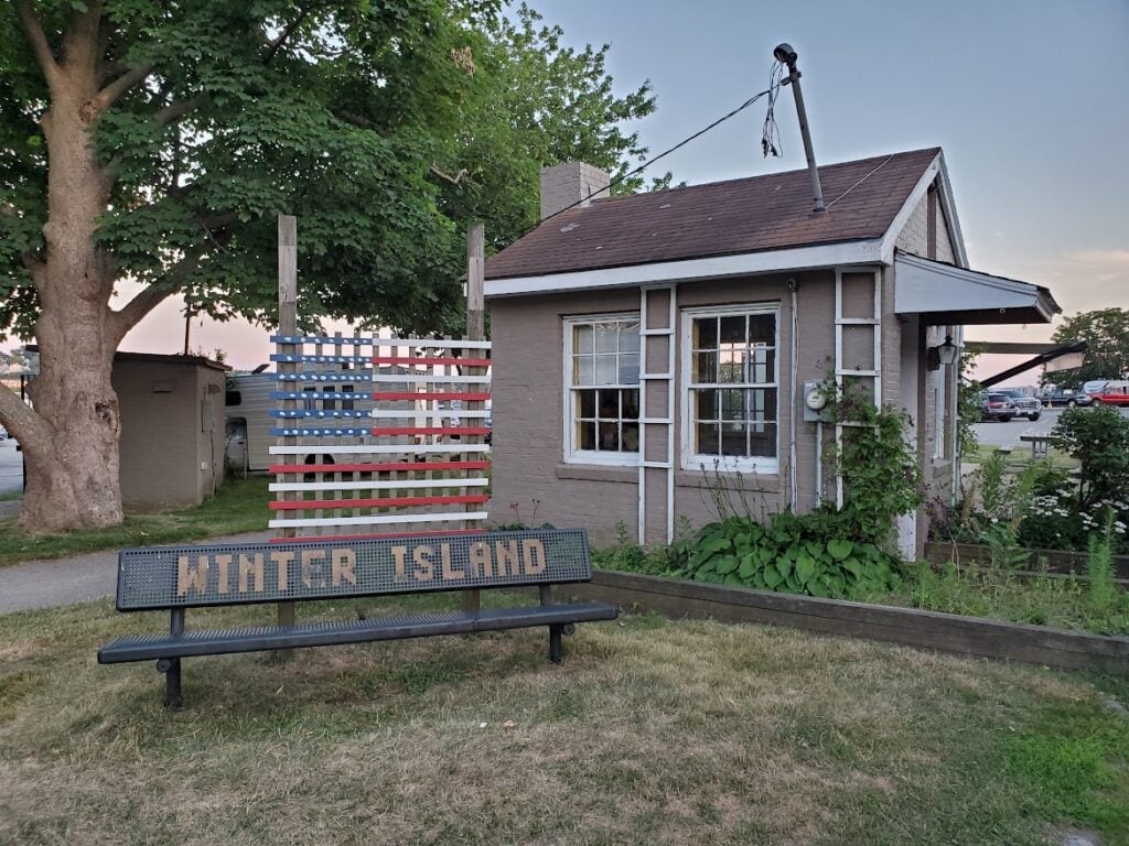 A shack with a wooden American flag next to it and a bench reading "Winter Island" in front.