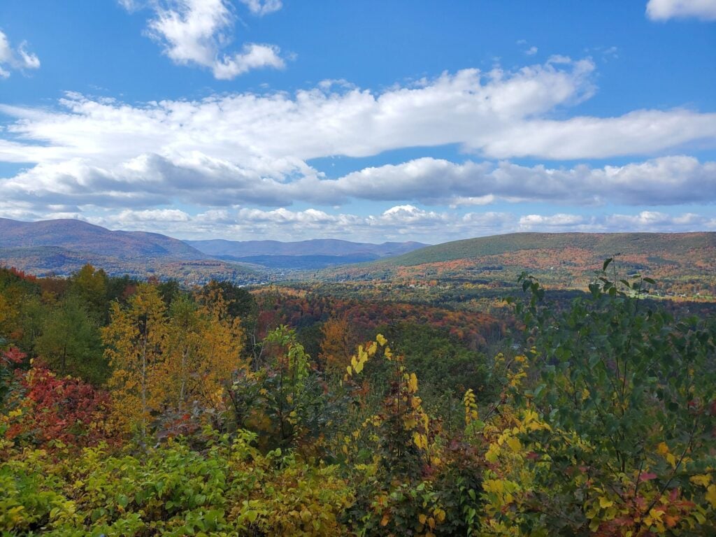 A scenic view of rolling hills and mountains covered in vibrant autumn foliage under a partly cloudy blue sky, much like the breathtaking vistas you’ll find at the 11 Spots to See Massachusetts Fall Foliage from High Above.