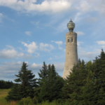 A tall, stone monument with a decorative top stands among green pine trees under a partly cloudy blue sky.
