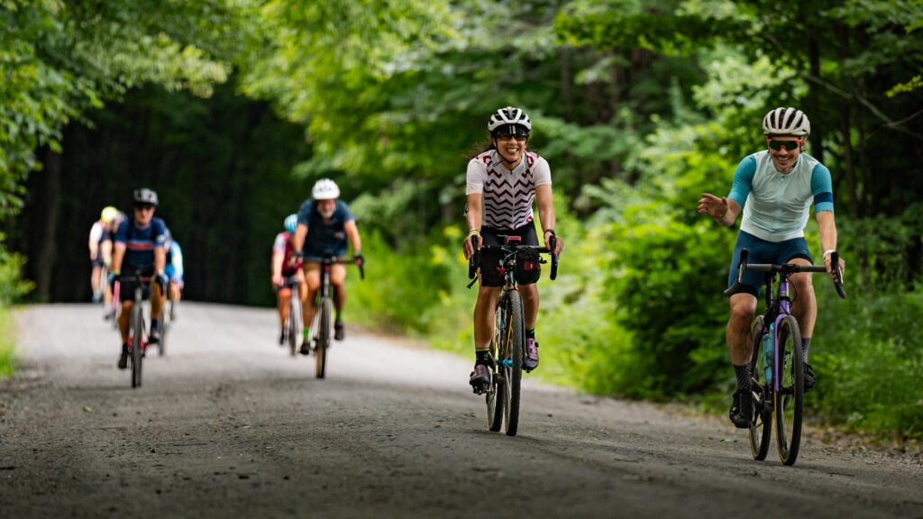 Cyclists travel down a dirt road surrounded by green trees.