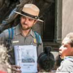 A park ranger in uniform holds a binder with an image and the text Battle of Ft. Wagner, SC, 1863, speaking to a group of children gathered around him outdoors.