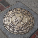 A bronze plaque set in the sidewalk marks the Freedom Trail in Boston, featuring a stylized torch and arrow with the words “The Freedom Trail Boston” engraved in a circular design.
