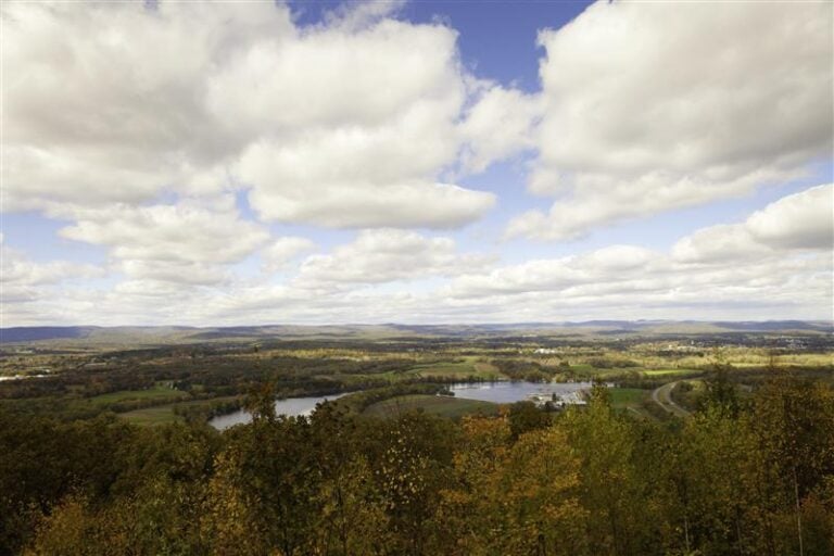 A scenic view of a rural landscape in Western Massachusetts captures 413 Day, with green fields, trees, a winding road, a small lake, and distant mountains under a partly cloudy sky.
