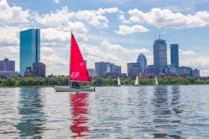 A sailboat with a red sail on the water in front of Boston's iconic skyline.