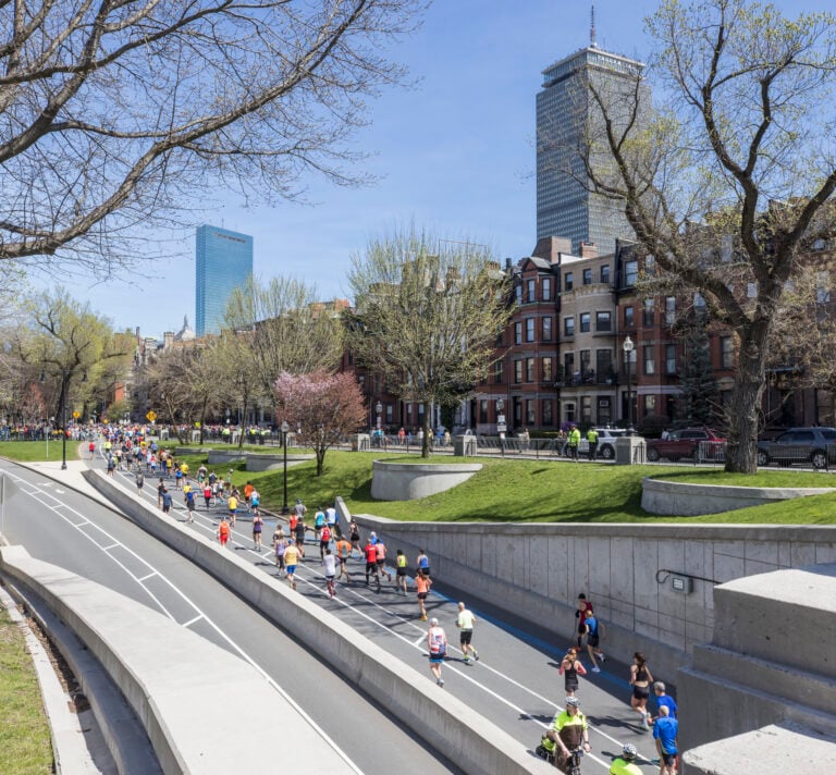 Runners participate in a marathon on a divided city street lined with trees and brownstone buildings, with tall skyscrapers visible in the background—capturing the spirit of the Boston Marathon Runner Guide on a clear, sunny day.