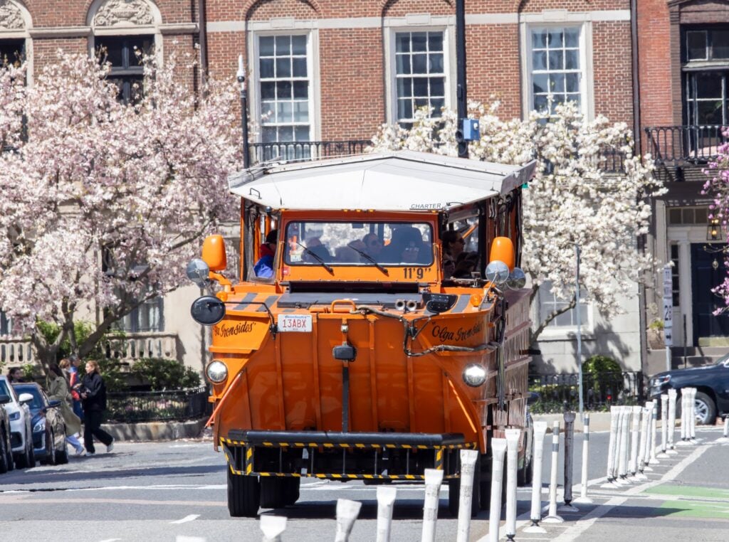 An orange amphibious tour bus drives down a city street lined with blooming trees and historic brick buildings on a sunny day, capturing the essence of An Unforgettable Summer in Massachusetts.