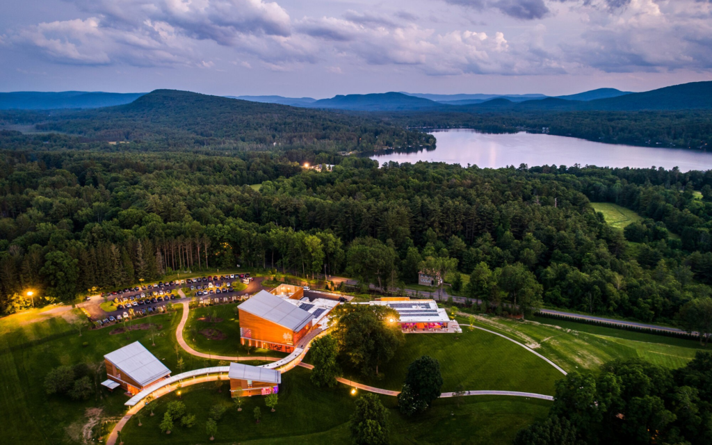 Aerial view of a modern building complex in one of the Best Towns in Western Massachusetts, surrounded by dense green forest, with a lake and mountains in the background. Winding pathways and illuminated areas promise summer fun near the buildings.