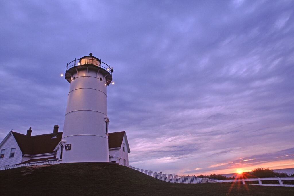 A white lighthouse stands on a grassy hill beside a house, its light shining at dusk. Celebrate National Lighthouse Day by admiring Massachusetts lighthouses, as clouds and hints of purple frame the orange sunset on the horizon.