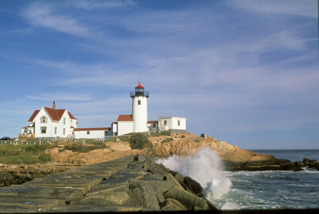 A white lighthouse with a red roof stands on rocky terrain beside the ocean, next to a white house. Waves crash against a stone pier in the foreground under a partly cloudy blue sky—perfect for celebrating National Lighthouse Day.