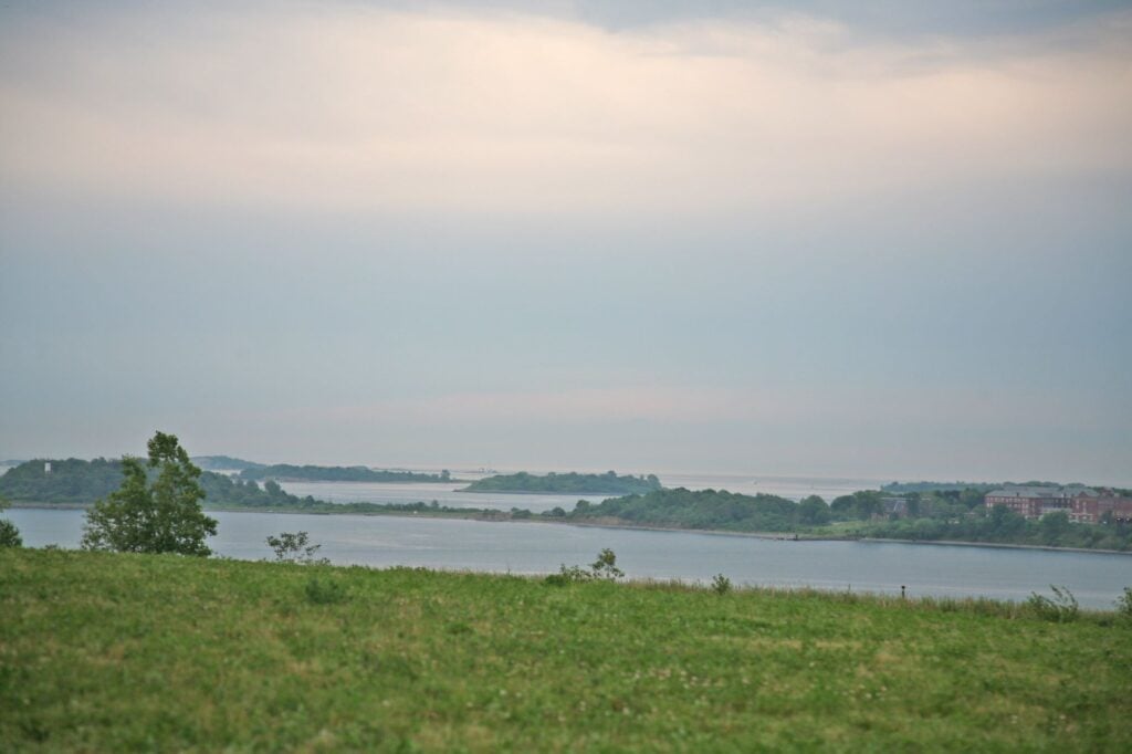 A grassy field stretches toward calm water, with small tree-covered islands in the distance under a cloudy sky at dusk—an inviting scene reminiscent of the best spring hikes in Massachusetts.
