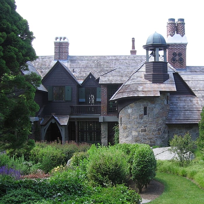 A large, rustic house in Massachusetts with stone walls, wooden siding, and multiple chimneys sits among lush green bushes and trees, featuring a round turret with a small cupola—perfect for LGBTQ+ travel enthusiasts seeking charm.