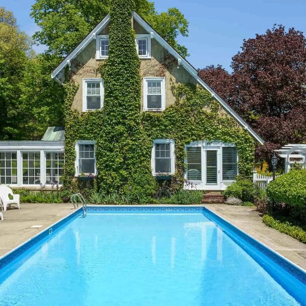 A charming house covered in green ivy stands behind a clear blue swimming pool—an inviting scene for your next trip planning, surrounded by lush trees and greenery under a bright blue sky.