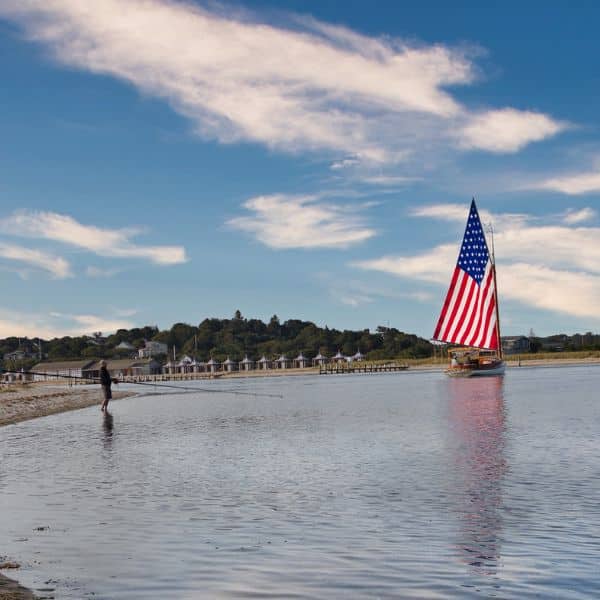 A sailboat with a sail resembling the American flag glides on calm water near the shore, inspiring creativity. A person stands at the water’s edge, as houses and trees dot the background beneath a blue sky.