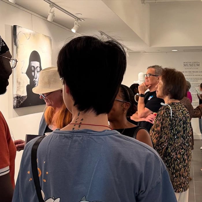 A diverse group of people in Massachusetts observe artwork in a brightly lit gallery. One painting depicts a face in shades of black and white. Visitors, including those on LGBTQ+ travel, engage in conversation and closely examine the art on display.