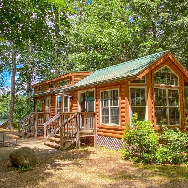 A cozy log cabin with large windows and a green metal roof sits among trees near a lake. Wooden steps lead to the front door, and a picnic table and boulders are nearby in the shaded, grassy yard—perfect to plan your trip outdoors.