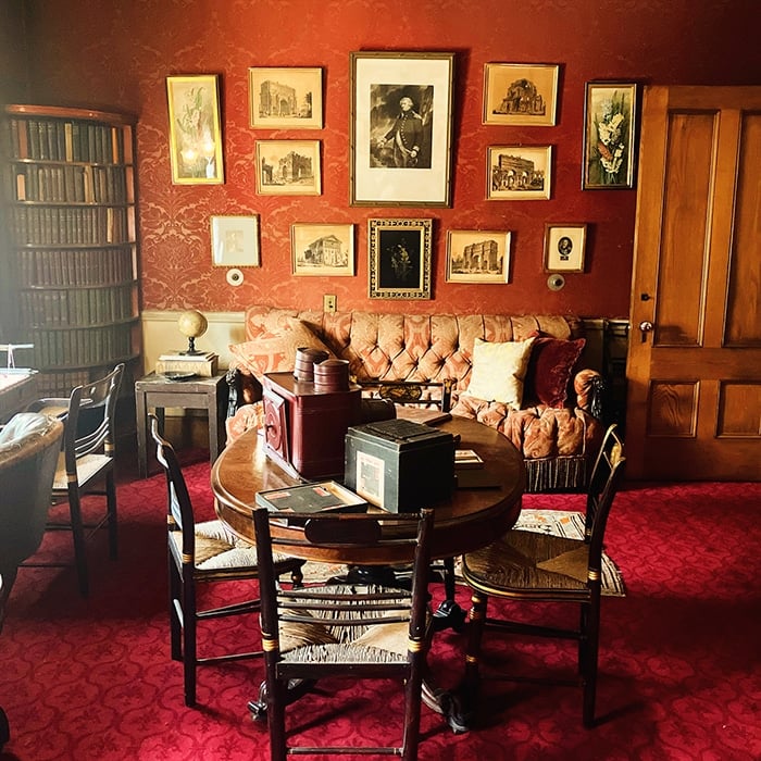 A cozy vintage study in Massachusetts with red wallpaper, a round table surrounded by chairs, a tan tufted sofa, bookshelves filled with books, and framed art and photos—an inviting space perfect for LGBTQ+ travel enthusiasts to unwind.