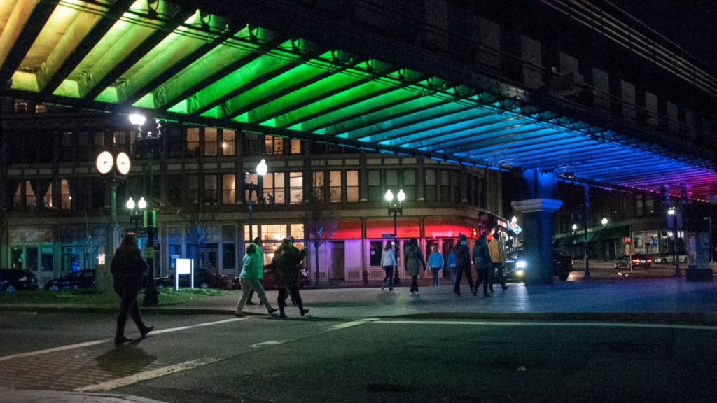 People cross a city street at night beneath a bridge in Lynn, the City of Firsts, illuminated with colorful lights in shades of green, blue, and red. Buildings with lit windows and street lamps glow in the background.