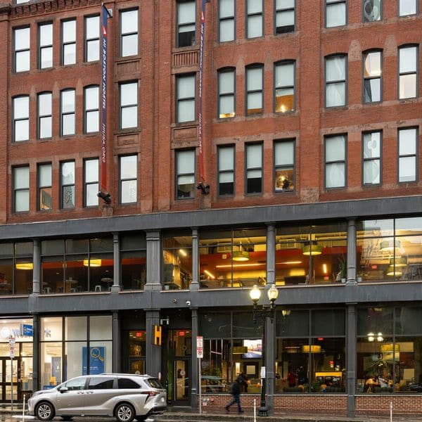 A red brick building with large windows on the first two floors and smaller ones above. Cars are parked on the wet street in front, and ground-level lights glow, hinting at businesses—perfect to explore as you plan your trip or for travel planning stops.