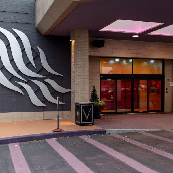 Modern building entrance with large abstract metal wall art, glass doors tinted red, a potted plant, and purple-striped pavement—perfect for snap-worthy trip planning stops. A cube with the letter V sits near the door.