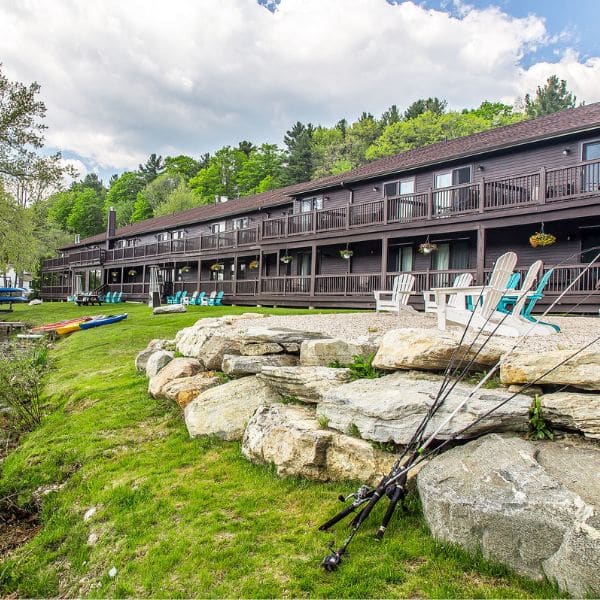 A rustic lodge with two stories and wooden balconies overlooks a grassy area dotted with fishing rods, Adirondack chairs, and kayaks—perfect for travel planning before exploring the calm lake surrounded by lush green trees under a partly cloudy sky.