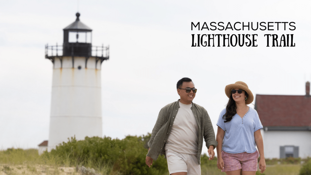 A couple walks on a sandy path with an iconic Massachusetts lighthouse behind them.