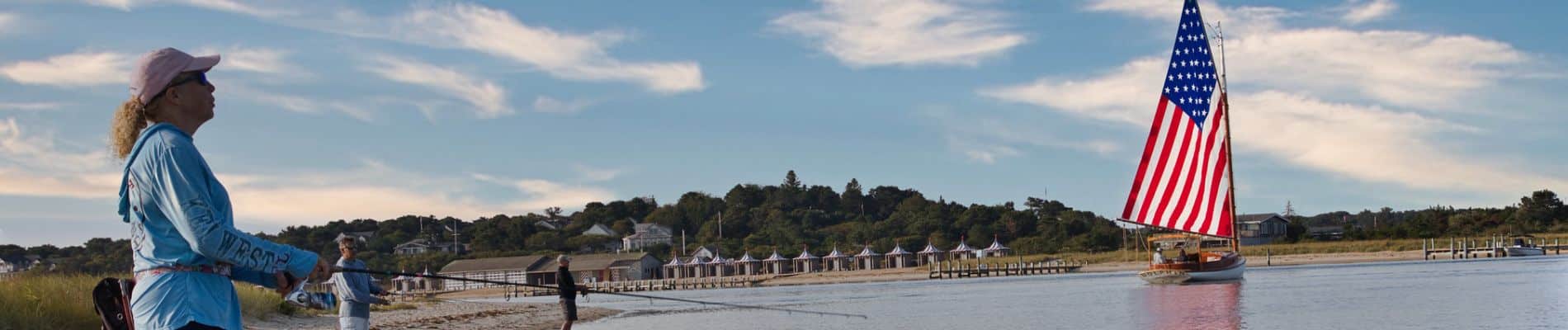 A person fishing on a sandy shore watches as a sailboat with an American flag sail glides across calm water, with houses and trees in the background under a blue sky.
