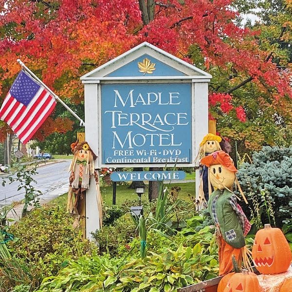 A blue sign for Maple Terrace Motel stands among colorful autumn foliage, perfect for trip planning. Surrounded by scarecrows and pumpkins, an American flag is displayed on the left side of the sign.