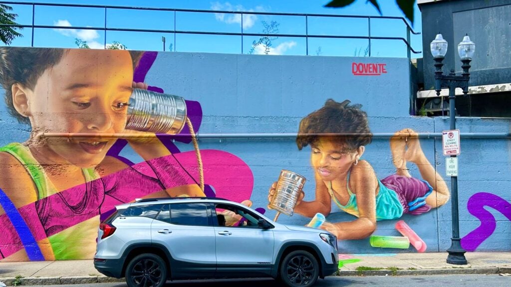 A car parked in front of a painted mural of 2 children playing.