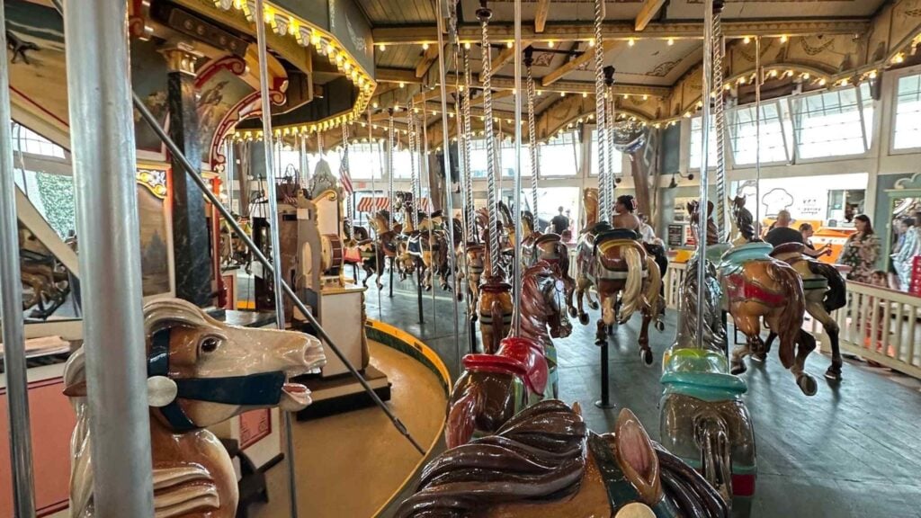 A brightly lit indoor carousel in Massachusetts with painted horses and animals, surrounded by people and large windows letting in daylight. The antique carousel features decorative lights and detailed artwork on its upper panels.