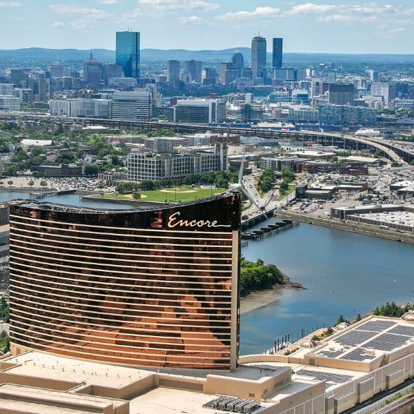 Aerial view of the Encore casino with its curved, copper-colored building in the foreground, set against the Boston city skyline—perfect inspiration as you plan your trip under a blue sky with scattered clouds.