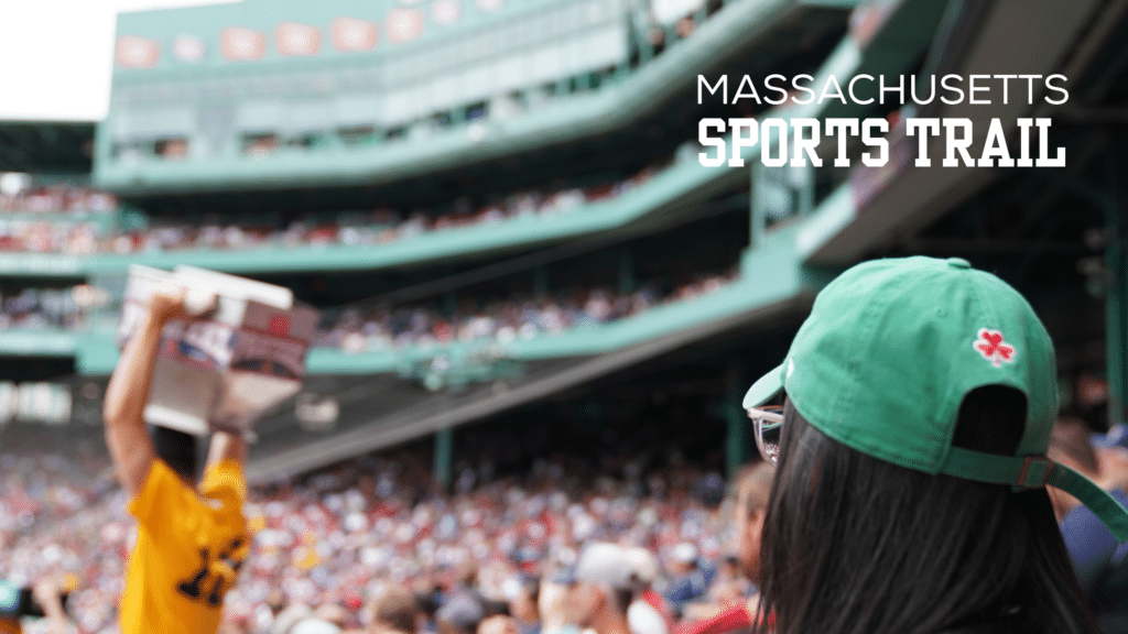 A lively crowd at a Massachusetts baseball stadium, featuring a person in a green cap and a fan holding a sign in the background. Text reads: Massachusetts Sports Trail.