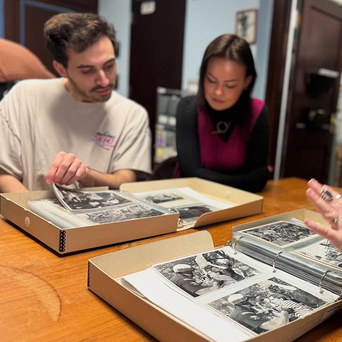 Two people sit at a table in Massachusetts, looking through binders filled with black and white photographs—perhaps planning LGBTQ+ travel. One points at a photo while the other turns a page, as a third hand reaches for another binder.