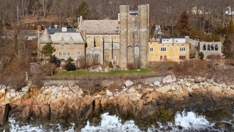 A large historic stone mansion, reminiscent of stately castles to visit north of Boston, sits atop a rocky shoreline amid leafless trees and a grassy lawn, with waves crashing against the rocks below.