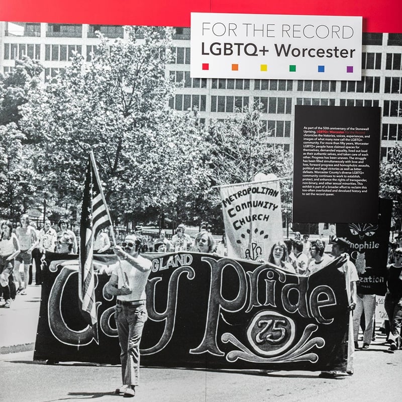Black-and-white photo of a group marching in a Gay Pride 75 parade, holding a large banner and American flag. The image, part of the FOR THE RECORD LGBTQ+ Worcester exhibit, highlights Massachusetts’ role in LGBTQ tourism.