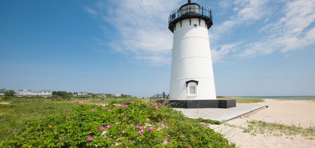 A white lighthouse stands near a sandy beach with green grass and pink flowers in the foreground, perfect for first-timer exploring on Martha’s Vineyard. Houses and clear blue sky are visible in the background.
