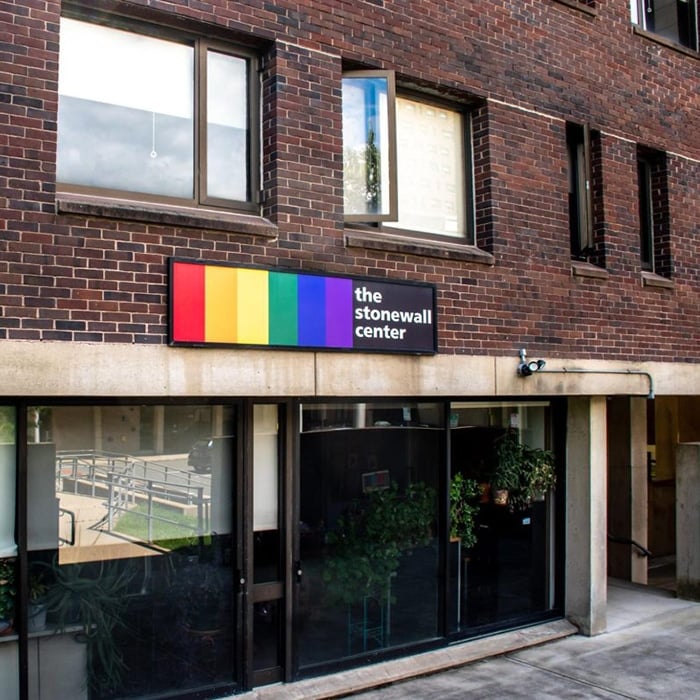 A brick building with large windows and a sign above one entrance reads The Stonewall Center beside vertical rainbow stripes, symbolizing LGBTQ+ pride. Green plants are visible inside—an iconic spot for LGBTQ+ travel in Massachusetts.