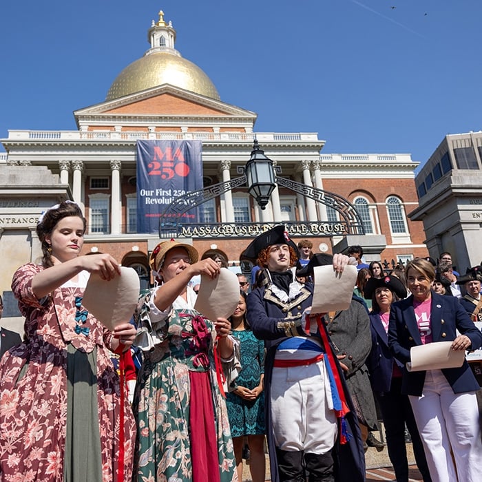 People in historical costumes tear papers during an outdoor event at the Massachusetts State House as media capture the moment. A crowd watches and a "MA 250" banner hangs on the building under a clear blue sky.