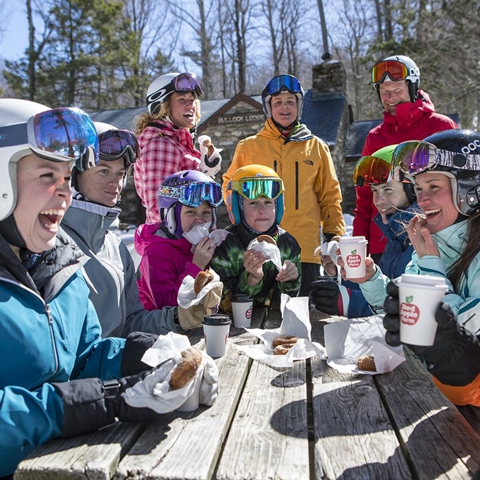 A group of people in winter gear and helmets enjoy hot drinks and pastries while sitting at a wooden picnic table outdoors, surrounded by trees, bright sunlight, and sharing industry resources.