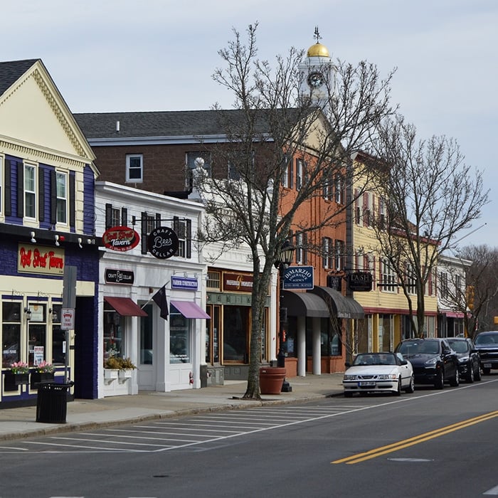 A small-town main street with colorful storefronts, parked cars along the road, leafless trees, and a building with a golden dome in the background offers a charming scene often used in media resources on a cloudy day.