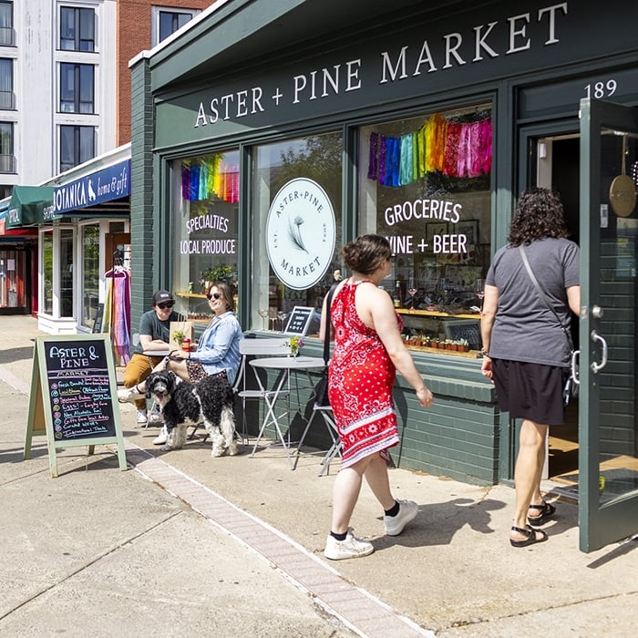 People enter and sit outside Aster + Pine Market, a grocery store with a green exterior. A dog sits beside a person at a table. Colorful ribbons hang above the shop’s window, and industry resources are listed on the sidewalk sign nearby.