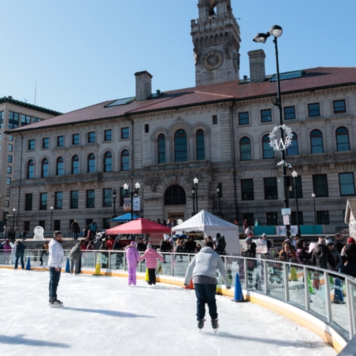 People ice skating on an outdoor rink in front of a large historic building with a clock tower, while others enjoy industry resources and media at nearby tents on a sunny day.