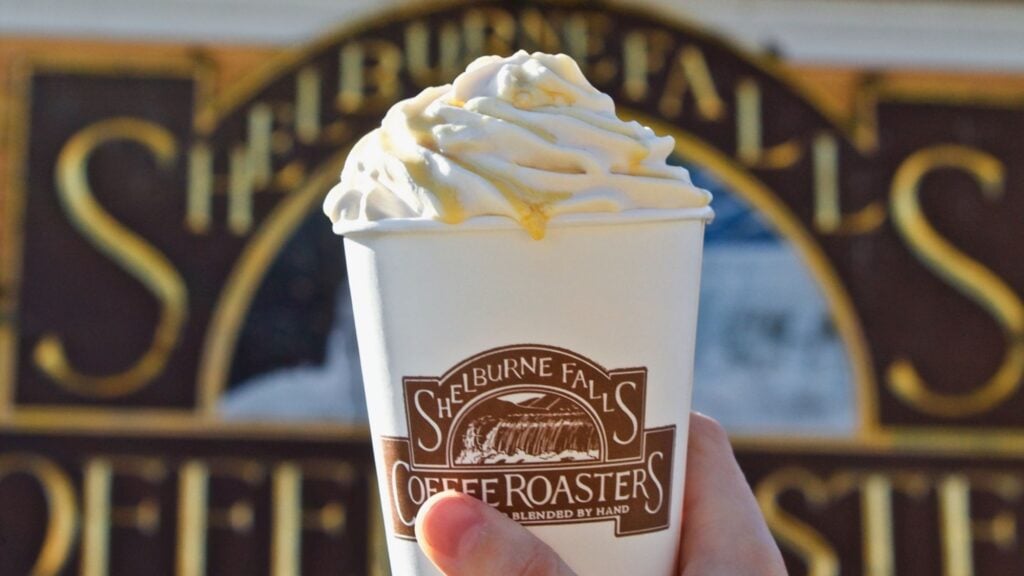 A paper cub with a Shelburne Falls Coffee Roasters logo, full of tasty treats with a dollop of whipped cream on top, with a Shelburne Falls Coffee Roasters sign in the background.