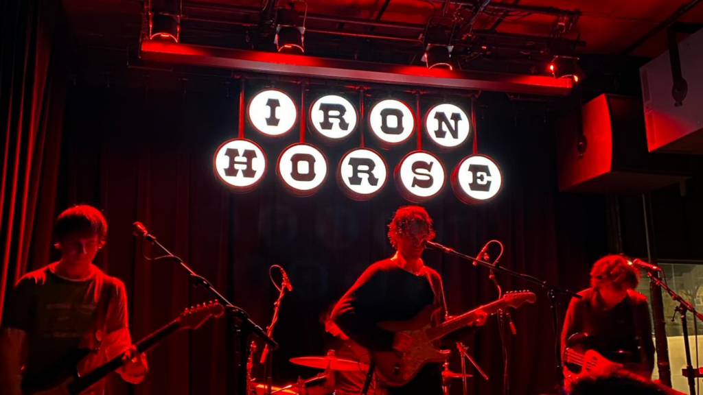 Two guitar players and a bassist perform on stage, bathed in red stage lights, underneath a lit white sign that reads "Iron Horse" at the Iron Horse performance venue in Northampton, Massachusetts.
