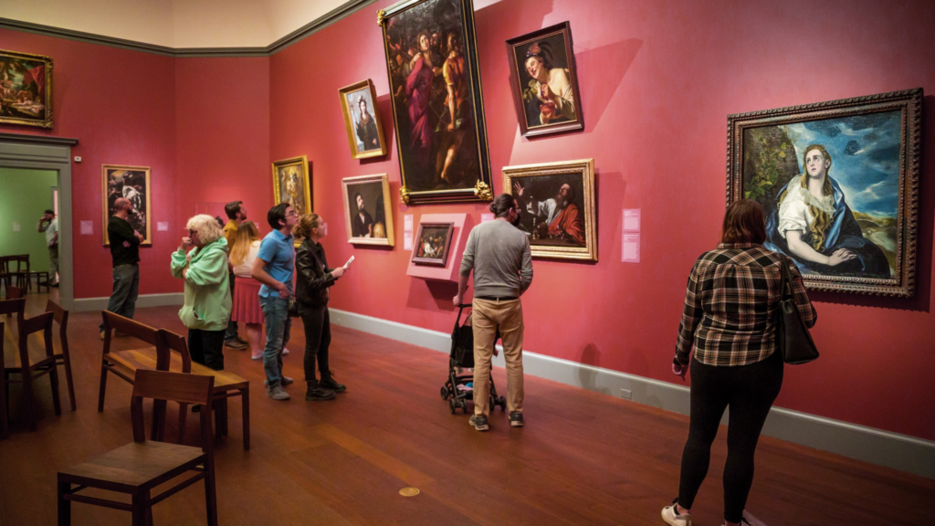 A group of museum-goers admire paintings in a red room.